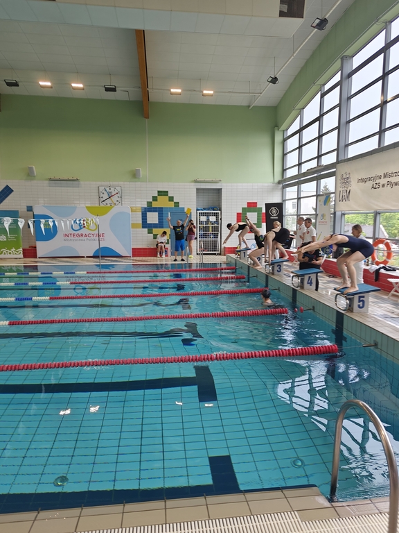 The interior of an indoor swimming pool during a swimming competition. Several swimmers in swimsuits stand on the starting posts, preparing to jump into the water. One of the swimmers is already in the middle of the jump.